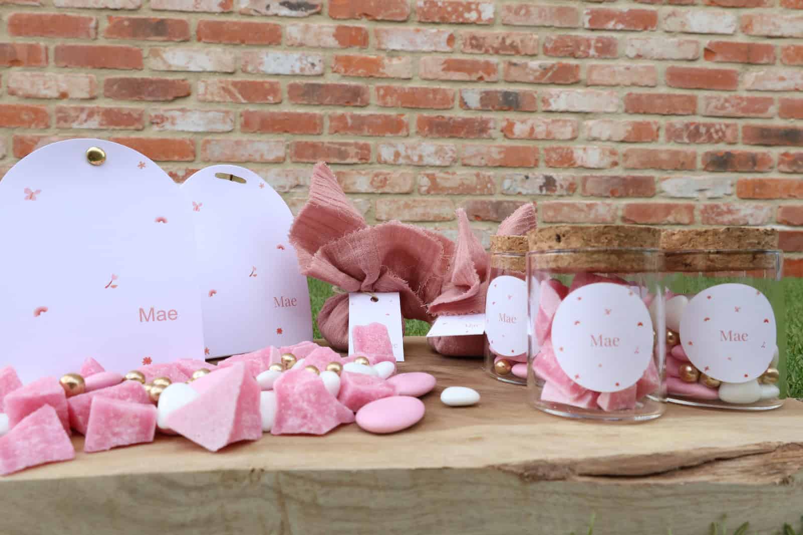Pastel roze snoepjes en bedankjes met naam "Mae" op een houten tafel, decoratie voor een verjaardagsfeest, buiten met een bakstenen muur op de achtergrond.